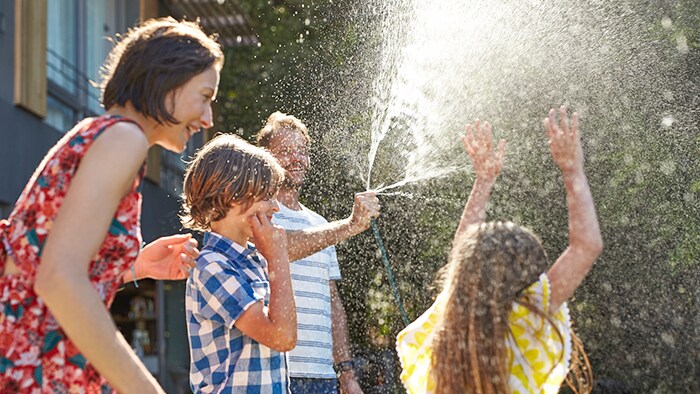 A father sprays a hose on a sunny day while his children run through the water and his wife smiles. 